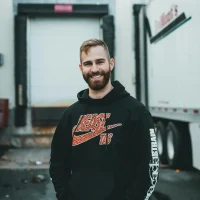 A smiling bearded man wearing a hoodie stands confidently outdoors by a truck.